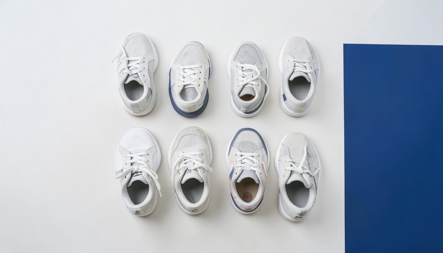 Clean overhead photograph of several sneakers arranged on a light neutral surface, white and grey tones with soft natural light